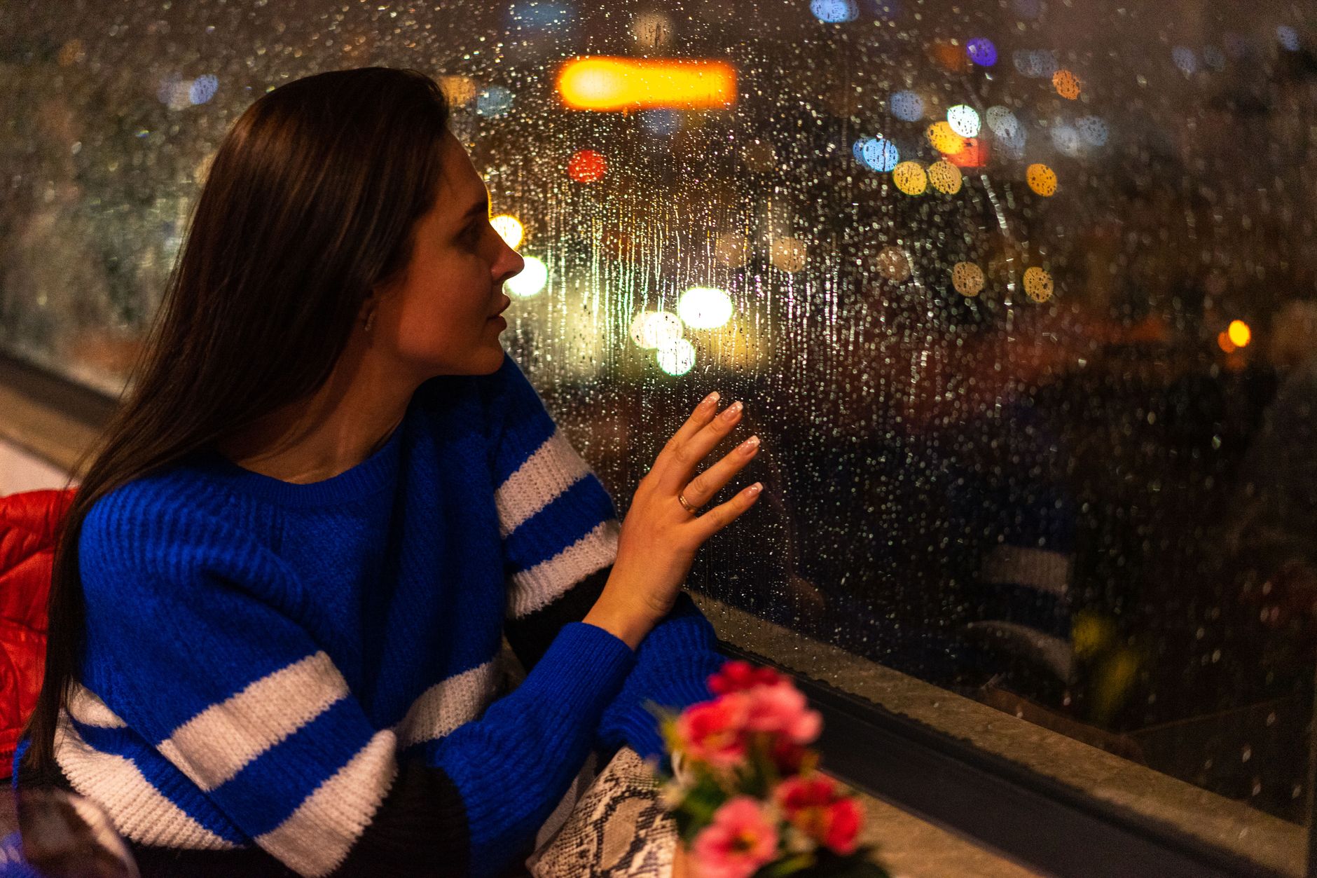 雨粒のついた窓に手を触れ夜の街明かりを見る女性