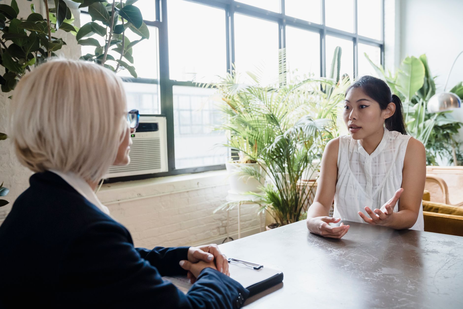 大きな窓と観葉植物のある室内でテーブル越しに話す二人の女性