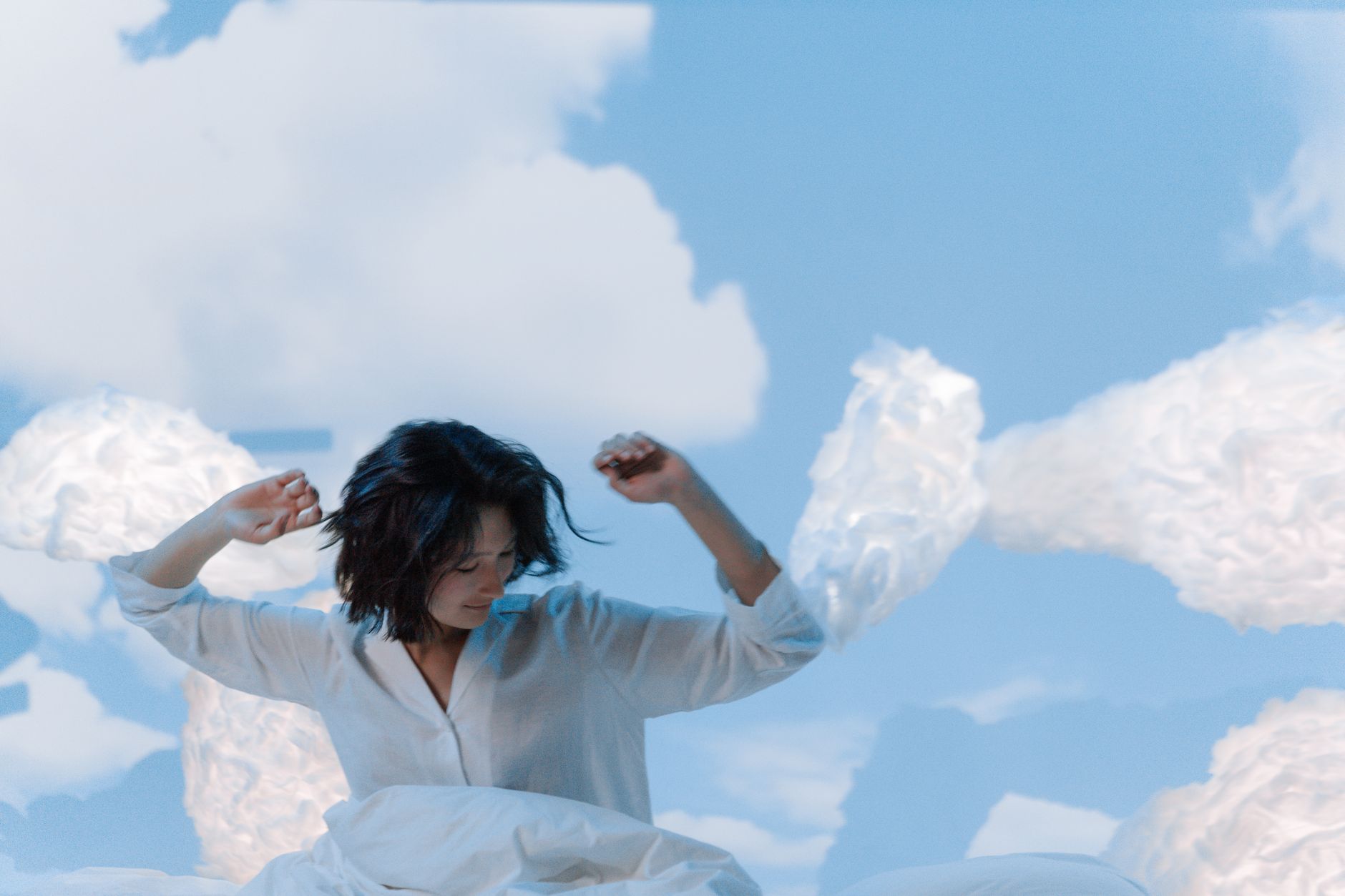 青空を背にベッドで伸びをする女性と綿雲のオブジェ