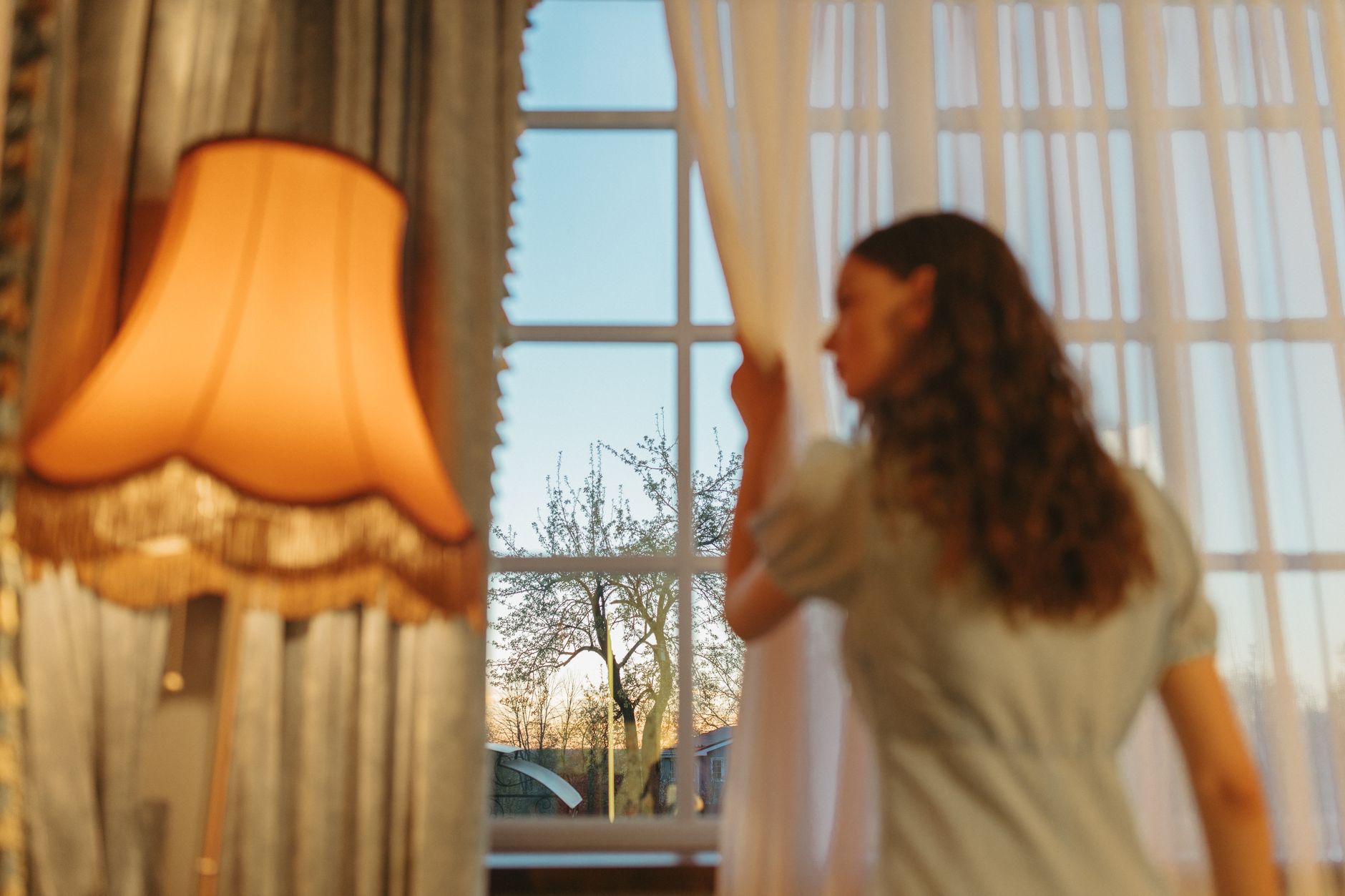 夕暮れのカーテンを開けて窓の外を見る女性