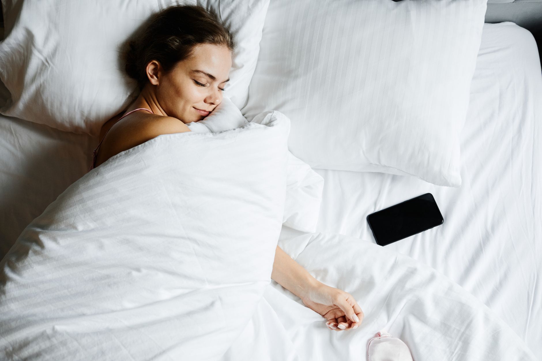 白いベッドで眠る女性とシーツの上のスマートフォンを真上から写した写真
