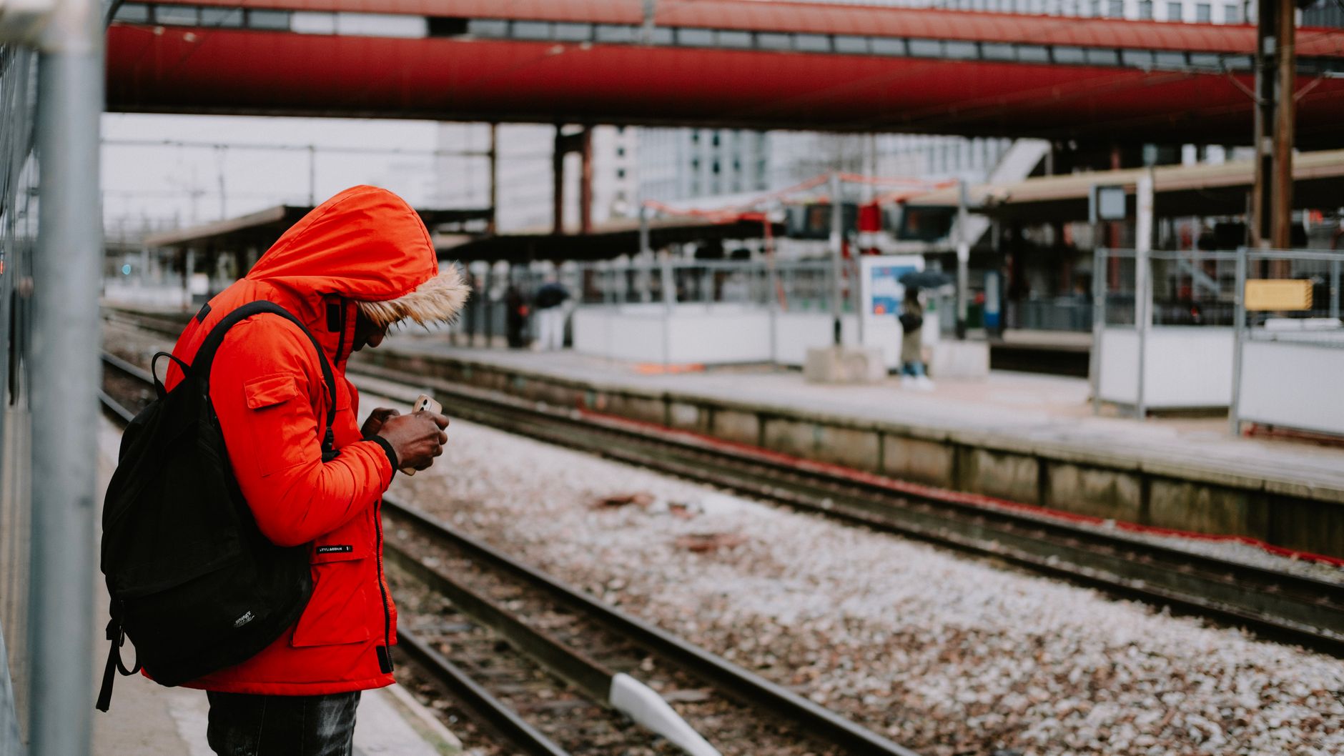 駅のホームでスマートフォンを確認する赤いジャケットの男性